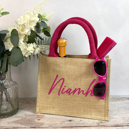 Jute bag with 'Niamh' branding, pink sunglasses, and a bottle on a wooden surface with flowers in the background.