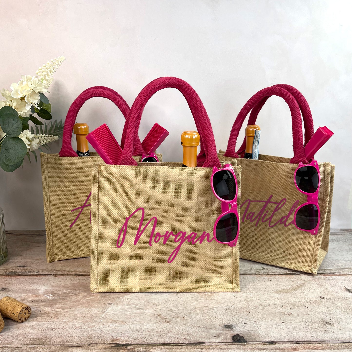 Three jute bags with pink handles and sunglasses, featuring personalized names, on a wooden surface.