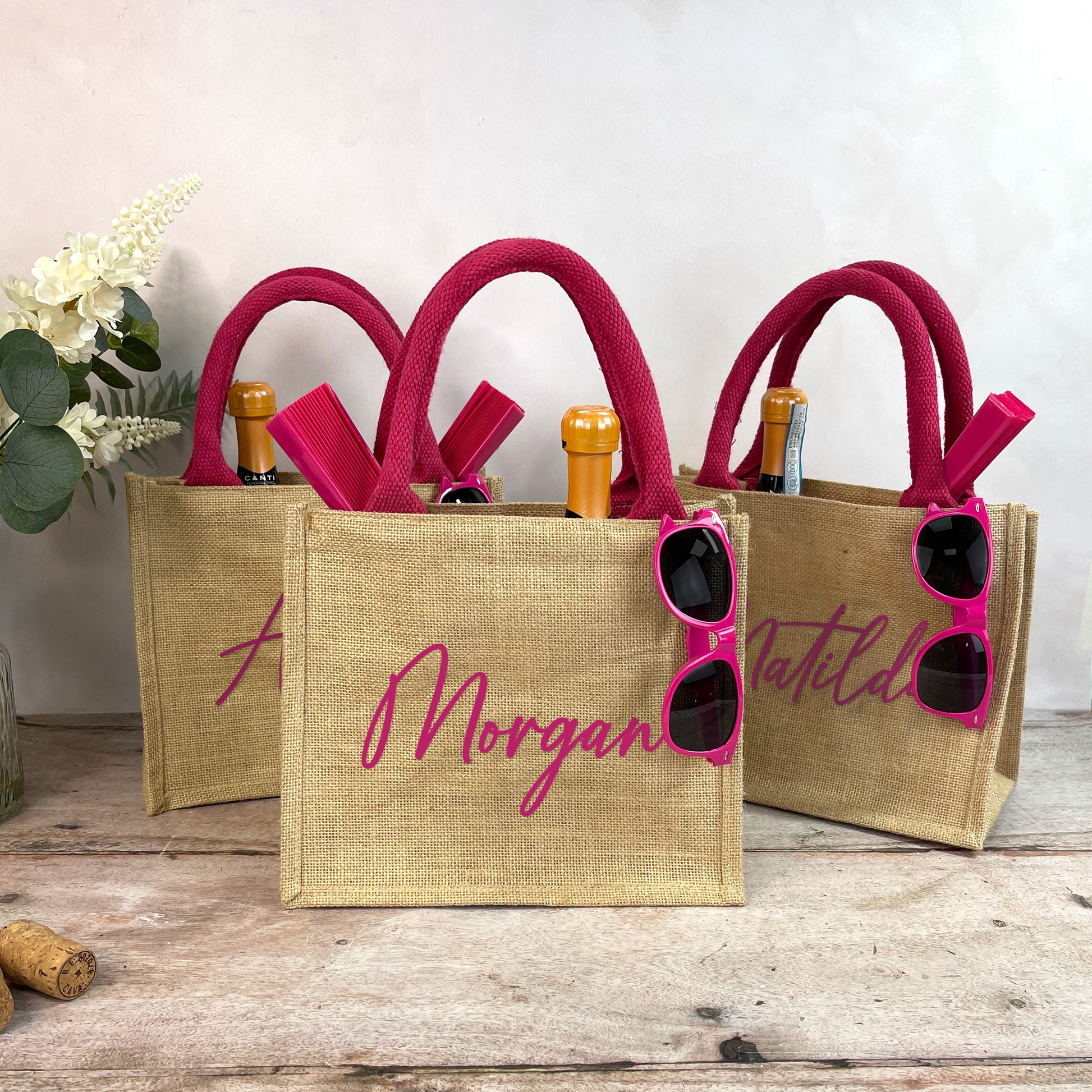 Three jute bags with pink handles and sunglasses, featuring personalized names, on a wooden surface.