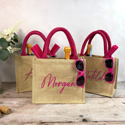Three jute bags with pink handles and sunglasses, featuring personalized names, on a wooden surface.