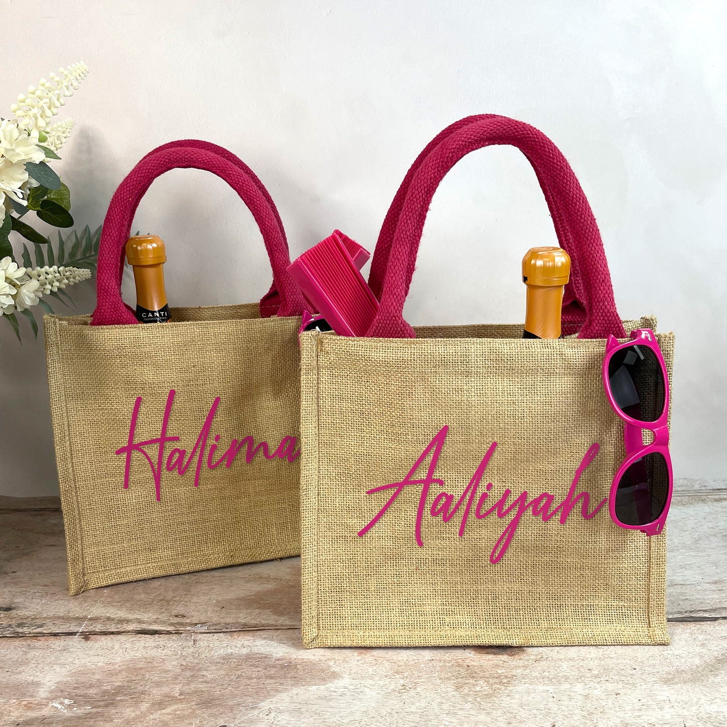 Two jute bags with pink handles and personalized names 'Halima' and 'Aaliyah', with sunglasses and a bottle on a wooden surface.