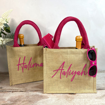 Two jute bags with pink handles and personalized names 'Halima' and 'Aaliyah', with sunglasses and a bottle on a wooden surface.