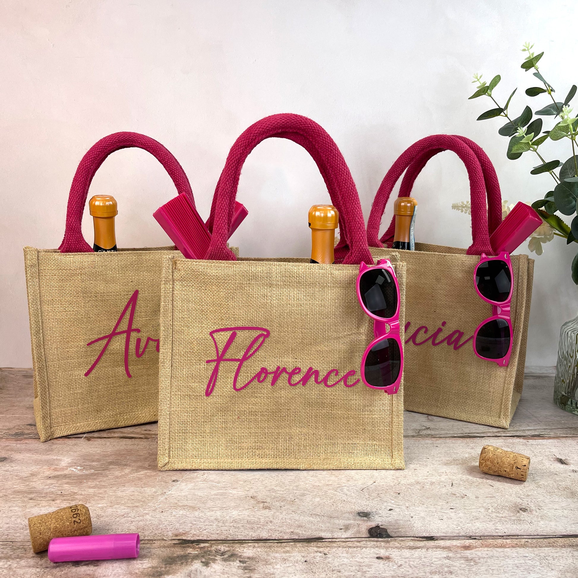 Three jute bags with pink handles and sunglasses, on a wooden surface with a light background.