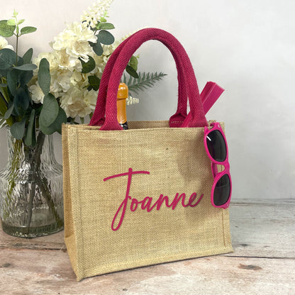 Jute bag with pink handles and sunglasses, featuring the name 'Joanne', on a wooden surface with flowers in the background.