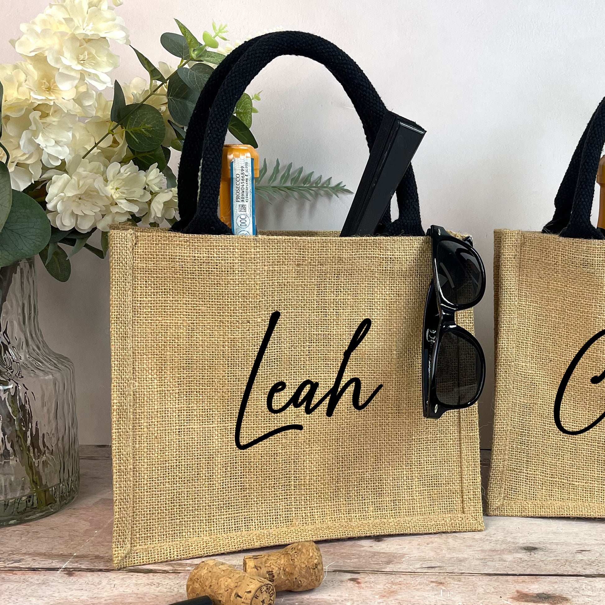 Jute bags with personalized names 'Leah' and 'C' on a wooden surface with flowers and a plant in the background.