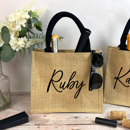 Two burlap bags with 'Ruby' and 'Katie' written on them, with sunglasses and a bottle inside, on a wooden surface.