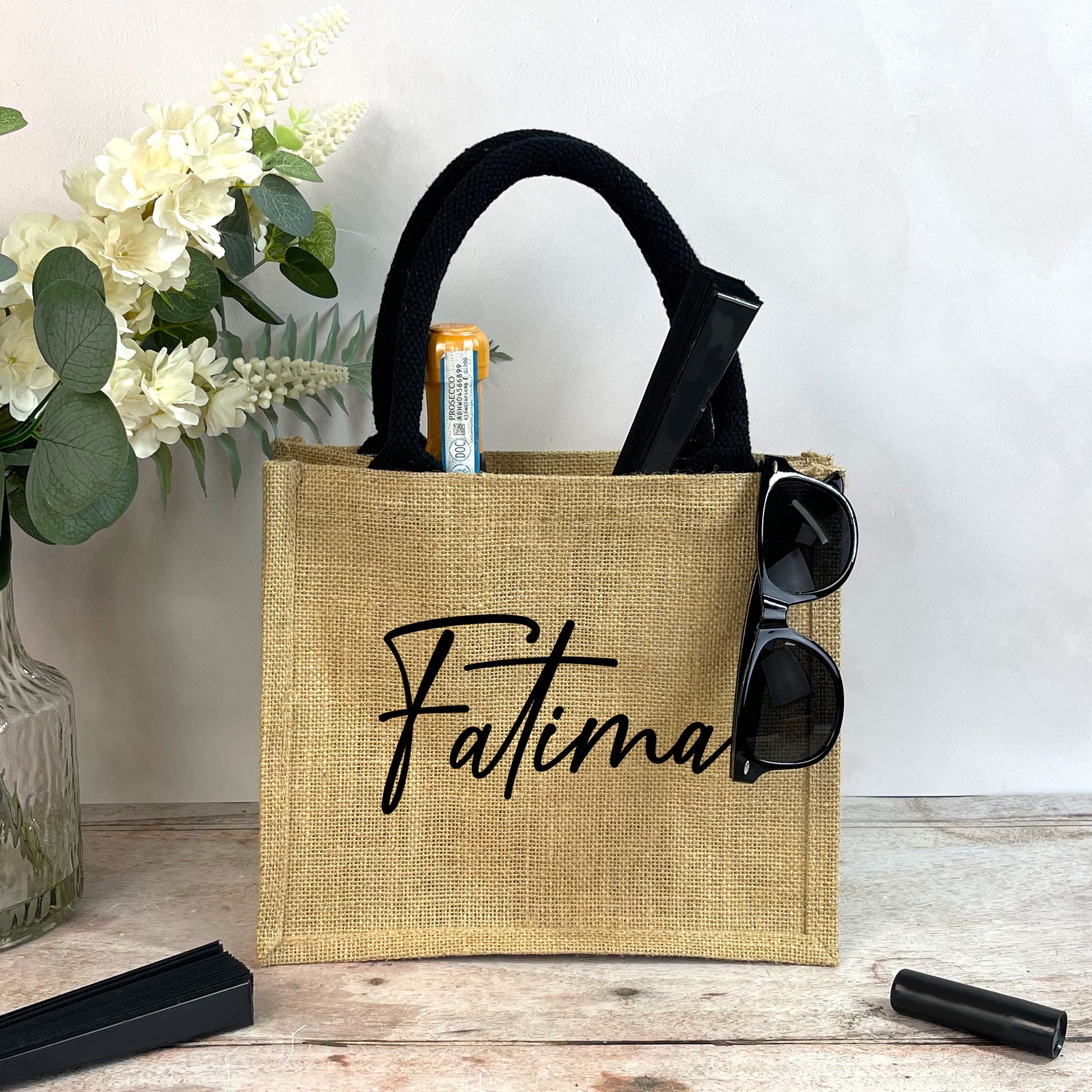 Jute bag with 'Fatima' branding on a wooden surface with flowers in the background
