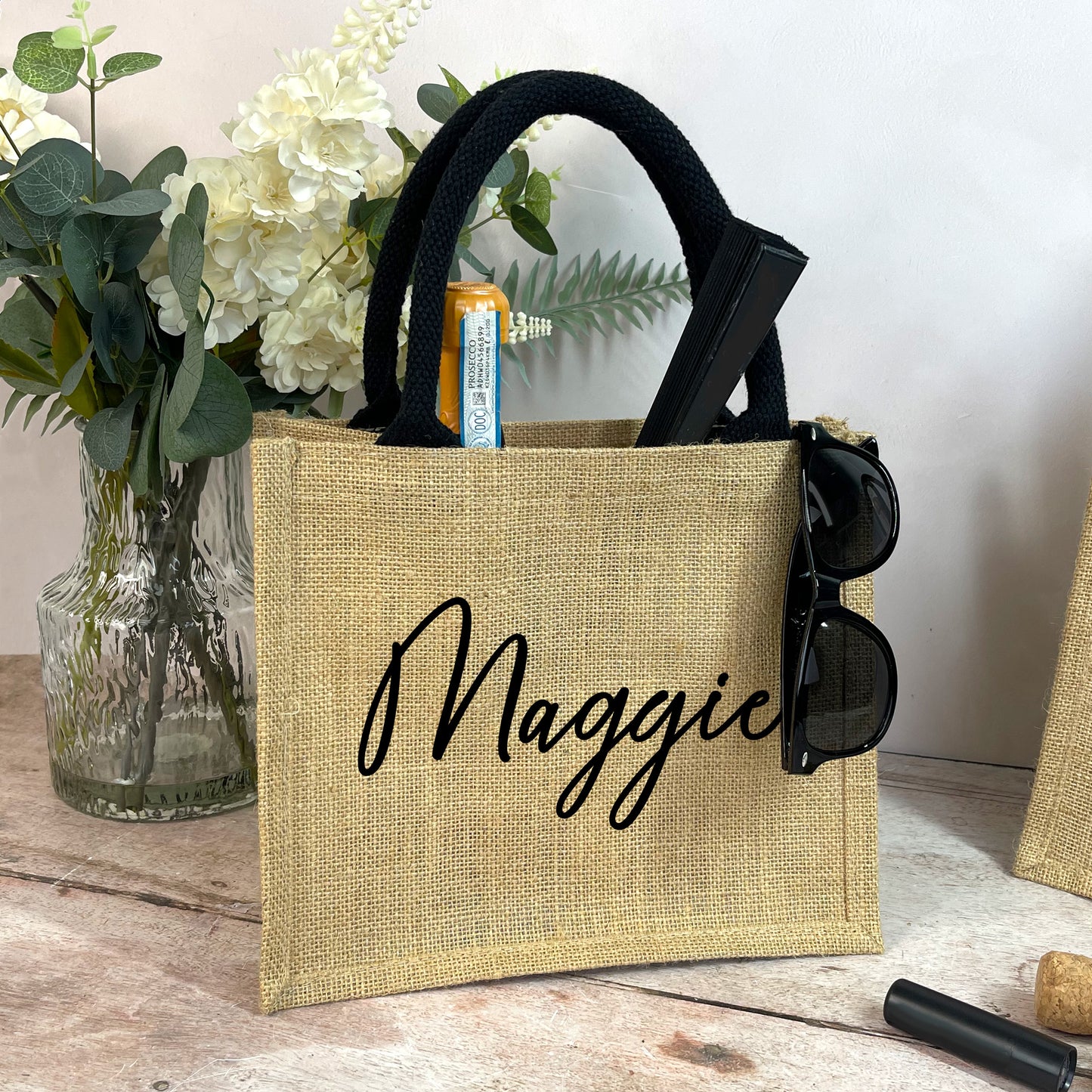 Beige tote bag with 'Maggie' printed on it, containing sunglasses and a bottle, on a wooden surface with flowers in the background.