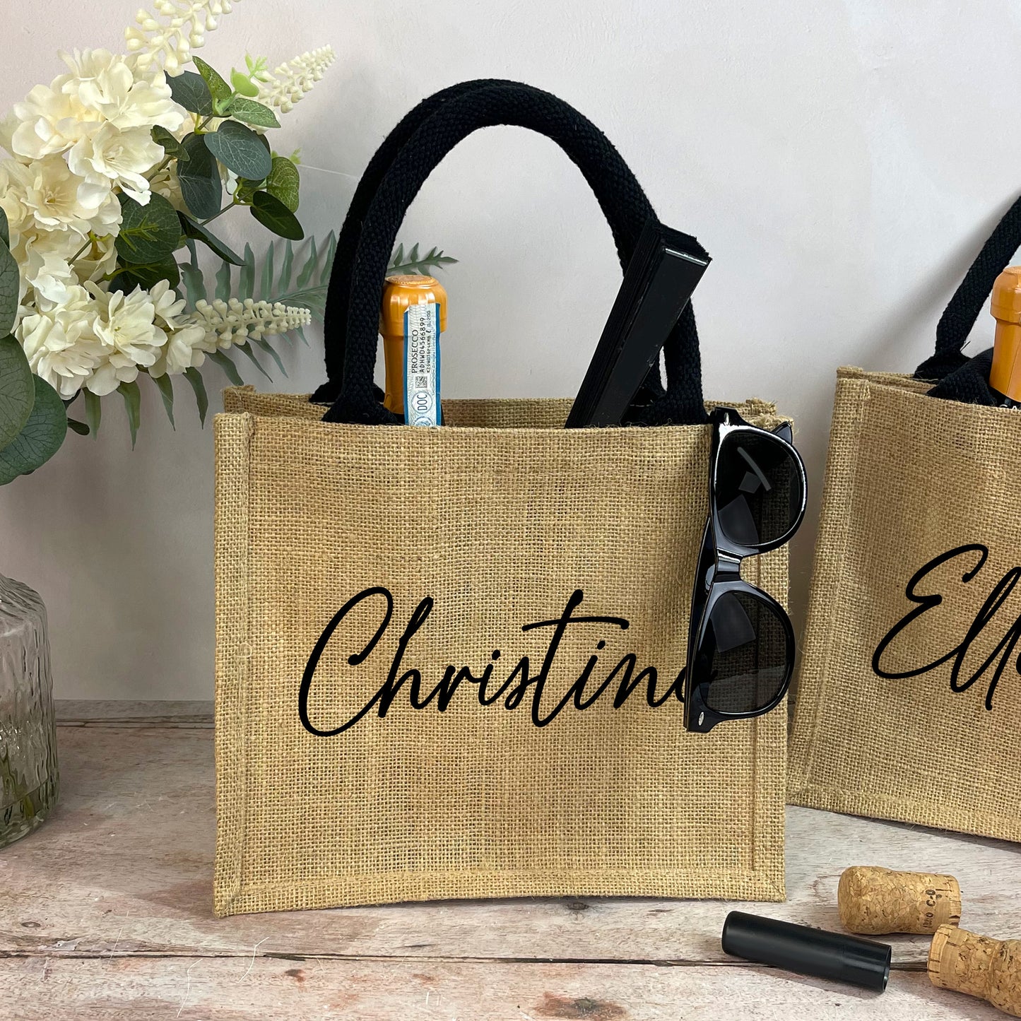 Two burlap bags with personalized names 'Christina' and 'Elle' on a wooden surface with flowers and accessories.