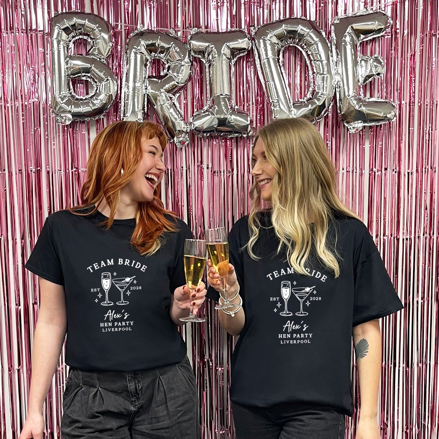 Two women standing in front of a pink metallic fringe backdrop with large silver 'BRIDE' balloons, wearing matching black graphic T-shirts that read 'TEAM BRIDE' and 'Alice's BFF Party Liverpool' with cocktail illustrations.