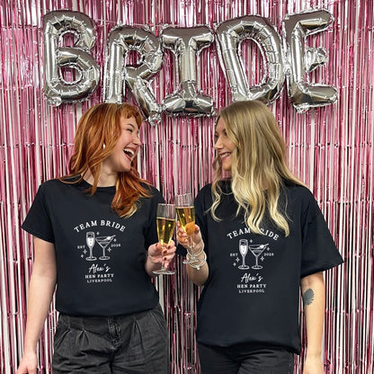 Two women standing in front of a pink metallic fringe backdrop with large silver 'BRIDE' balloons, wearing matching black graphic T-shirts that read 'TEAM BRIDE' and 'Alice's BFF Party Liverpool' with cocktail illustrations.
