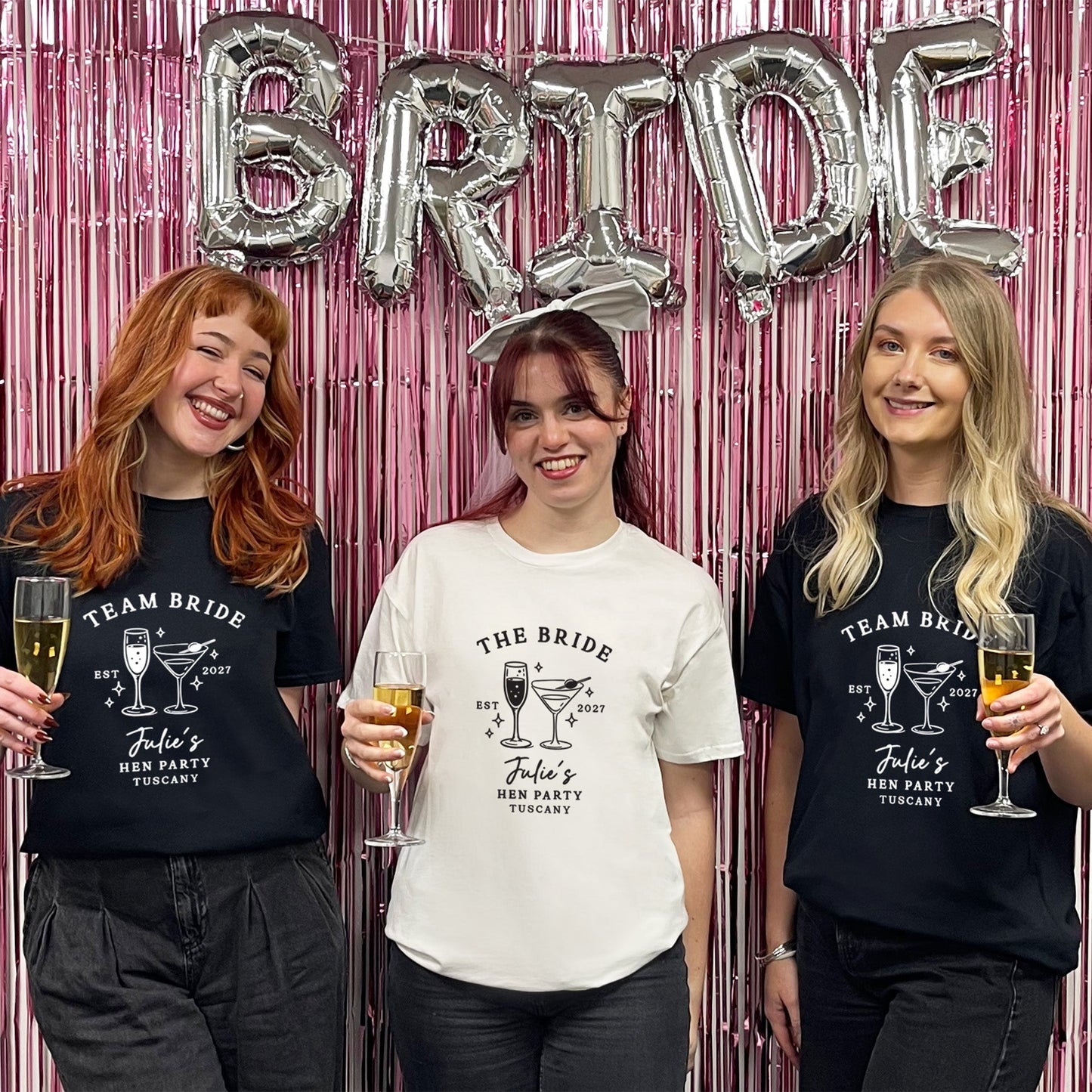 Three women standing together in front of a pink metallic fringe backdrop with silver foil balloons spelling BRIDE. Two women wear black graphic T-shirts reading TEAM BRIDE / John's Bachelorette Party T-Shirts with cocktail illustrations, and the center woman wears a white graphic T-shirt reading THE BRIDE / John's Bachelorette Party T-Shirt with cocktail illustrations.