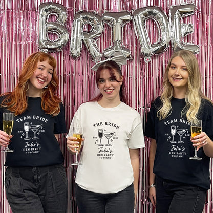 Three women standing together in front of a pink metallic fringe backdrop with silver foil balloons spelling BRIDE. Two women wear black graphic T-shirts reading TEAM BRIDE / John's Bachelorette Party T-Shirts with cocktail illustrations, and the center woman wears a white graphic T-shirt reading THE BRIDE / John's Bachelorette Party T-Shirt with cocktail illustrations.