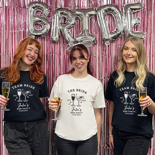 Three women standing together in front of a pink metallic fringe backdrop with silver foil balloons spelling BRIDE. Two women wear black graphic T-shirts reading TEAM BRIDE / John's Bachelorette Party T-Shirts with cocktail illustrations, and the center woman wears a white graphic T-shirt reading THE BRIDE / John's Bachelorette Party T-Shirt with cocktail illustrations.