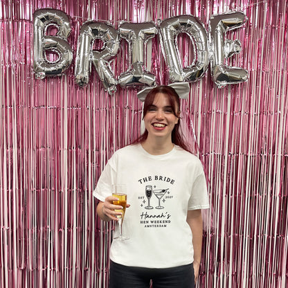 A woman stands in front of a pink metallic fringe backdrop with large silver 'BRIDE' balloons behind her, holding a glass of champagne. She is wearing a white graphic T-shirt that reads 'THE BRIDE' with cocktail illustrations and 'Hannah's BEE WEEKEND AMSTERDAM'.
