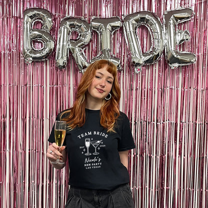 A woman with red hair stands in front of a pink metallic tinsel backdrop with large silver foil balloons spelling BRIDE. She is wearing a black graphic T-shirt that reads 'TEAM BRIDE' with cocktail illustrations and 'Nicole's Bachelorette Los Angeles,' and she holds a glass of champagne.