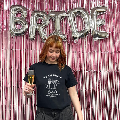A woman with red hair stands in front of a pink metallic tinsel backdrop with large silver 'BRIDE' balloons, wearing a black graphic T-shirt that reads 'TEAM BRIDE' and 'Cinema's Best Weekends Makers' with cocktail illustrations.