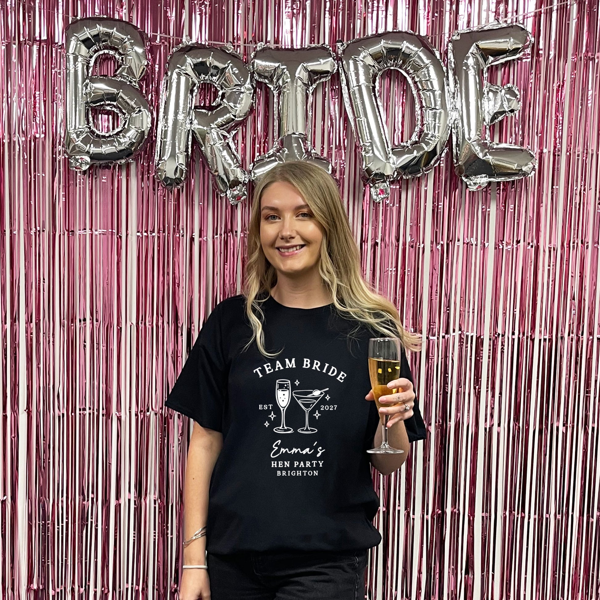 A woman stands in front of a pink metallic tinsel backdrop with large silver foil balloons spelling BRIDE. She is wearing a black graphic T-shirt that reads 'TEAM BRIDE Emma's Brides Party Brighton' with cocktail illustrations, and she holds a glass of champagne.