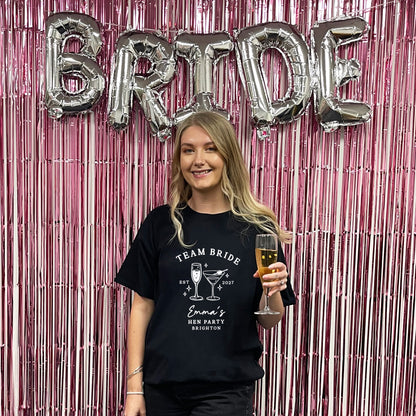 A woman stands in front of a pink metallic tinsel backdrop with large silver foil balloons spelling BRIDE. She is wearing a black graphic T-shirt that reads 'TEAM BRIDE Emma's Brides Party Brighton' with cocktail illustrations, and she holds a glass of champagne.