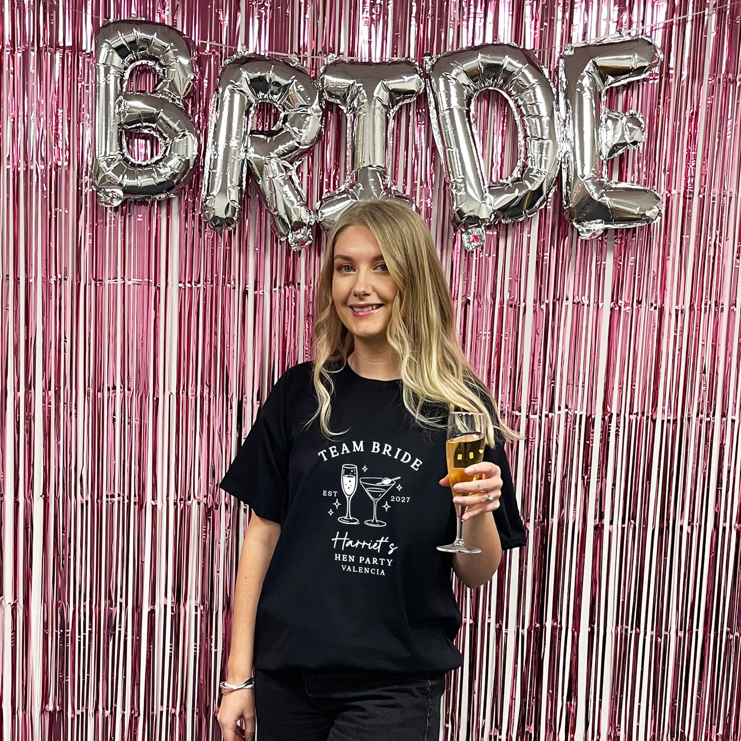 A woman stands in front of a pink metallic fringe backdrop with large silver foil balloons spelling BRIDE. She is wearing a black graphic T-shirt that reads 'TEAM BRIDE' with cocktail illustrations and text, and she holds a glass of champagne.