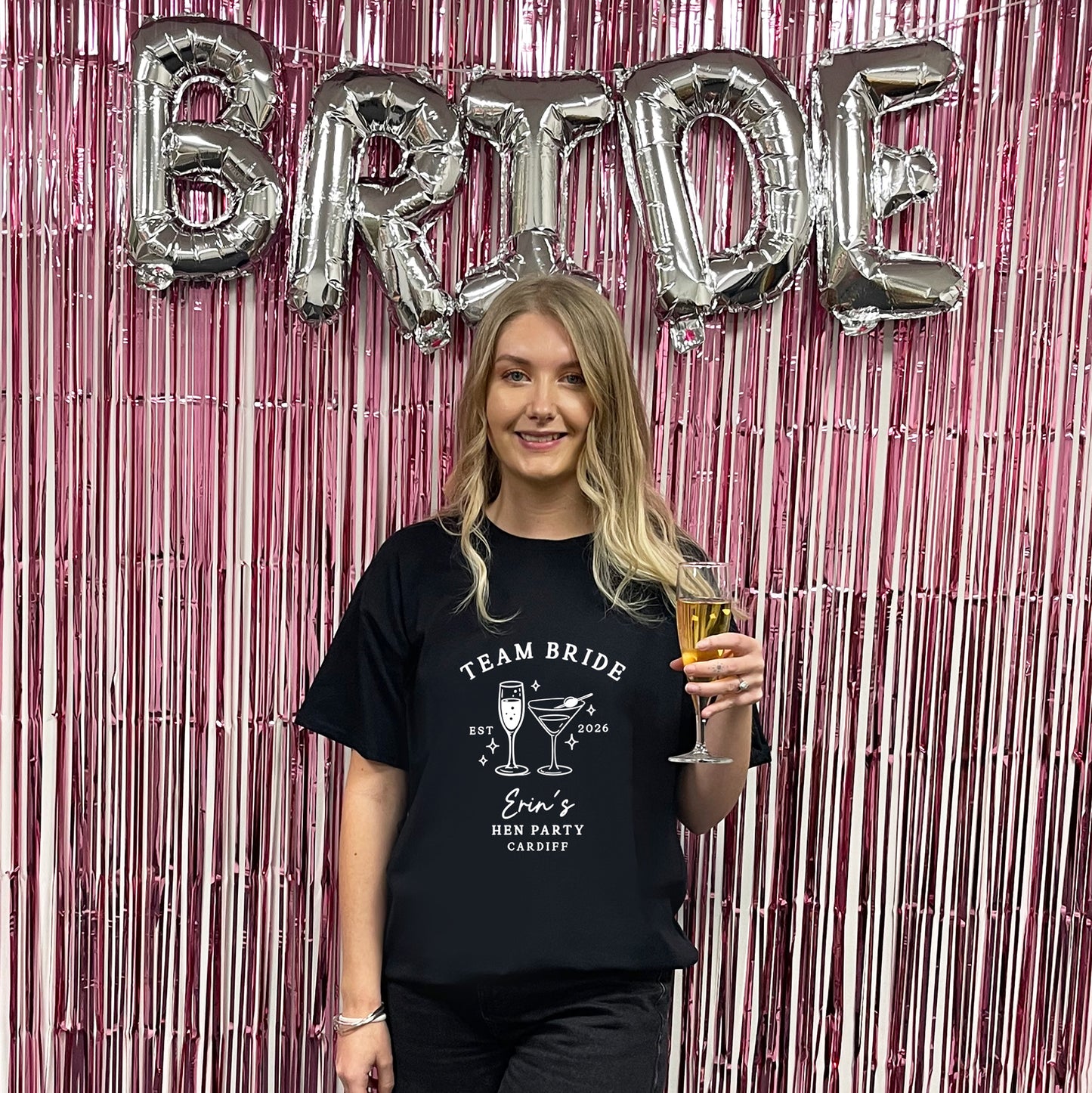 A woman stands in front of a pink metallic tinsel backdrop with large silver foil balloons spelling BRIDE. She is wearing a black graphic T-shirt that reads 'TEAM BRIDE' with cocktail illustrations and 'Bride's Best Party Cabbagg.