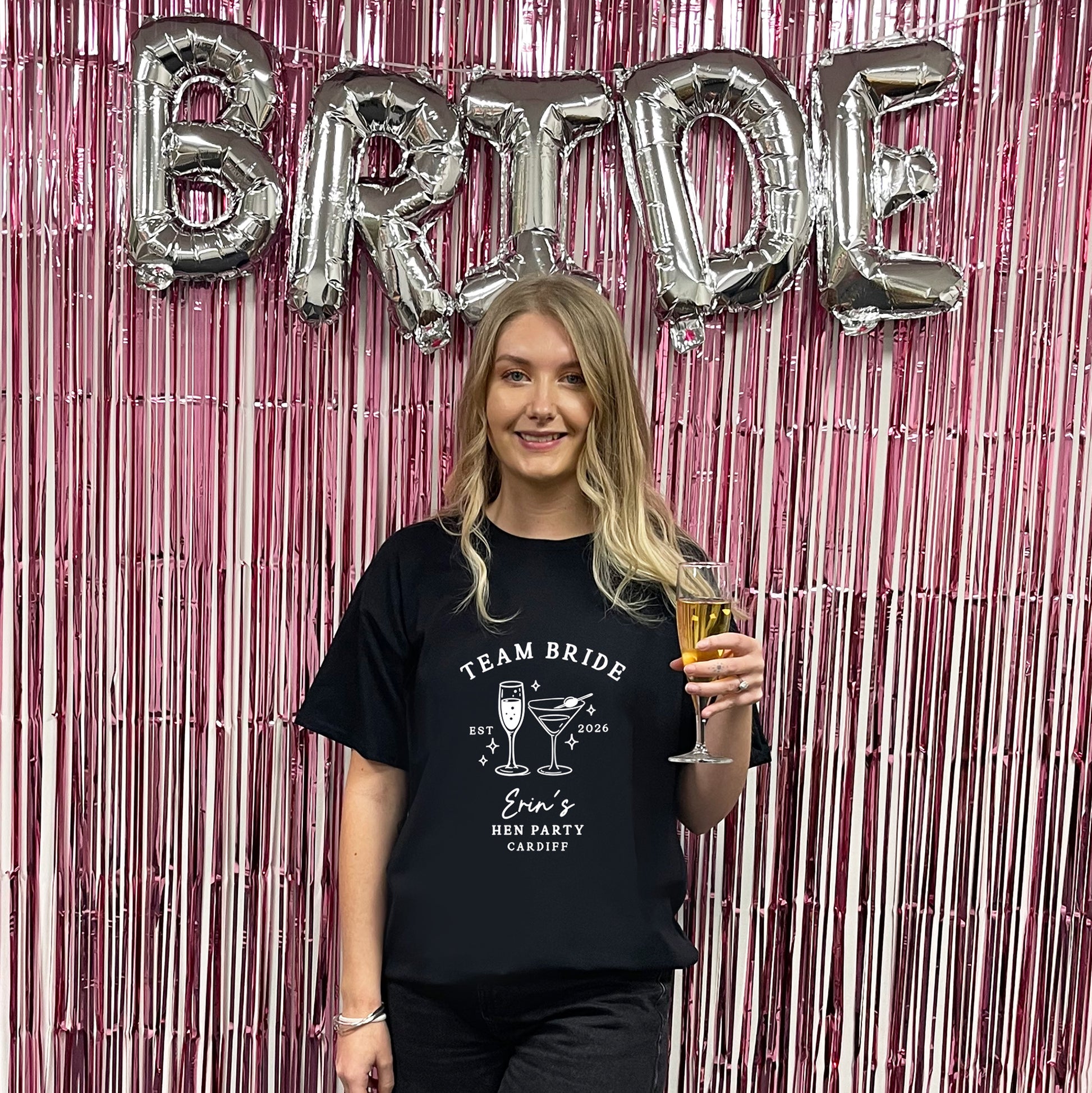 A woman stands in front of a pink metallic tinsel backdrop with large silver foil balloons spelling BRIDE. She is wearing a black graphic T-shirt that reads 'TEAM BRIDE' with cocktail illustrations and 'Bride's Best Party Cabbagg.