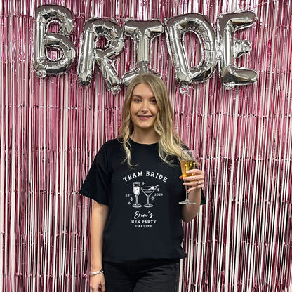 A woman stands in front of a pink metallic tinsel backdrop with large silver foil balloons spelling BRIDE. She is wearing a black graphic T-shirt that reads 'TEAM BRIDE' with cocktail illustrations and 'Bride's Best Party Cabbagg.