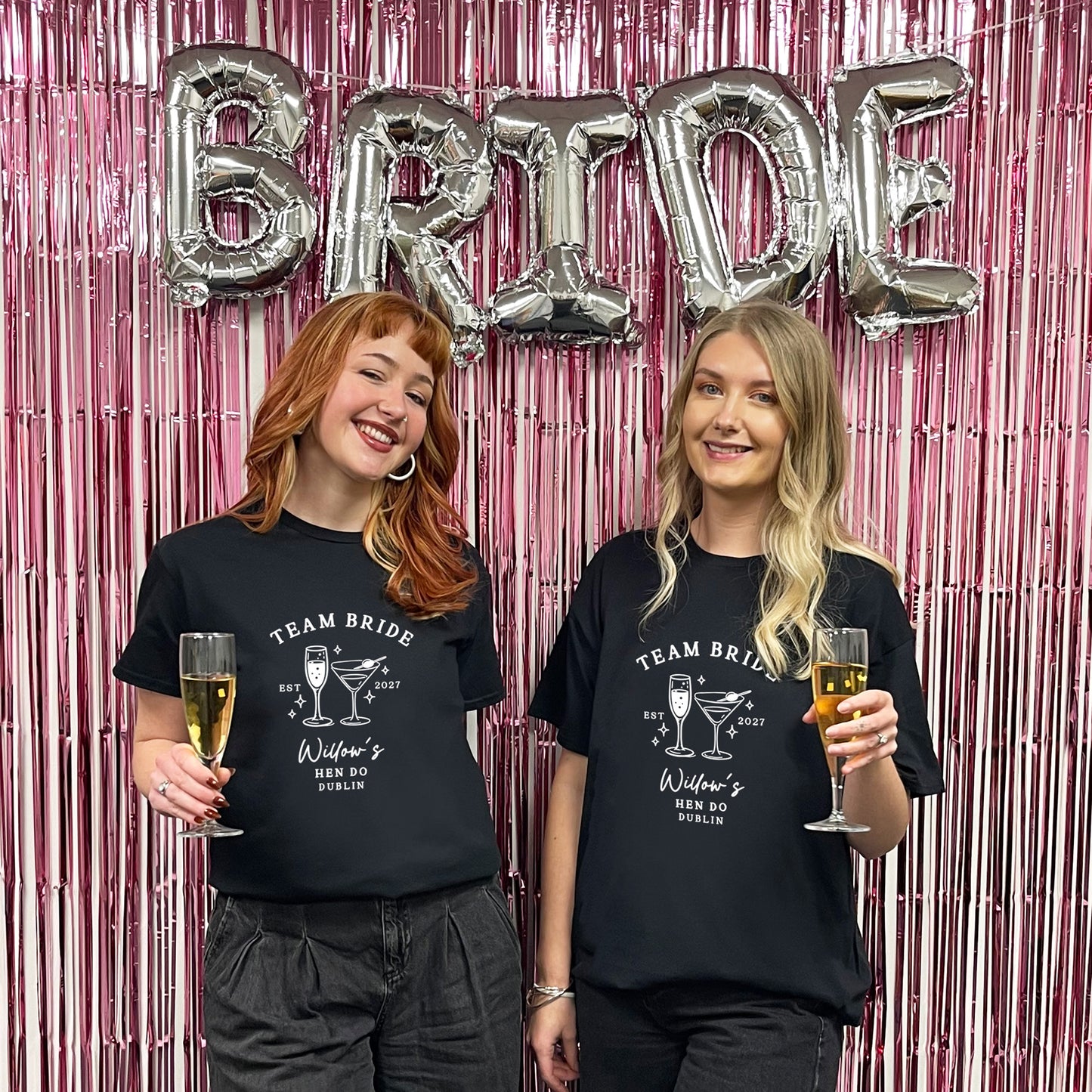 Two women standing in front of a pink tinsel backdrop with silver foil balloons spelling BRIDE, wearing matching black graphic T-shirts that read TEAM BRIDE with a cocktail illustration and text.