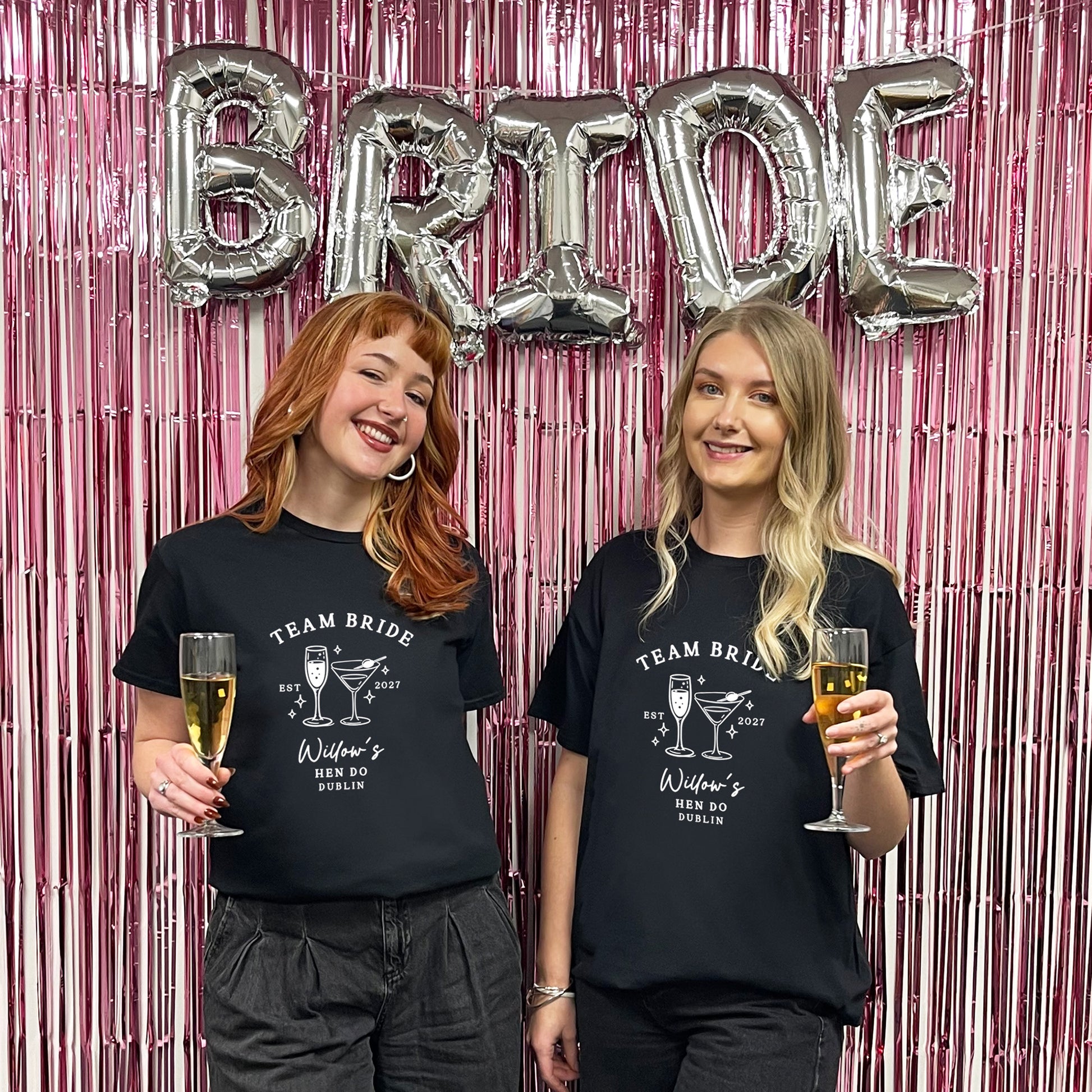 Two women standing in front of a pink tinsel backdrop with silver foil balloons spelling BRIDE, wearing matching black graphic T-shirts that read TEAM BRIDE with a cocktail illustration and text.