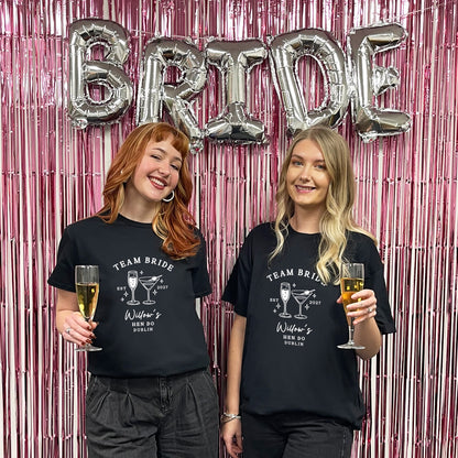 Two women standing in front of a pink tinsel backdrop with silver foil balloons spelling BRIDE, wearing matching black graphic T-shirts that read TEAM BRIDE with a cocktail illustration and text.