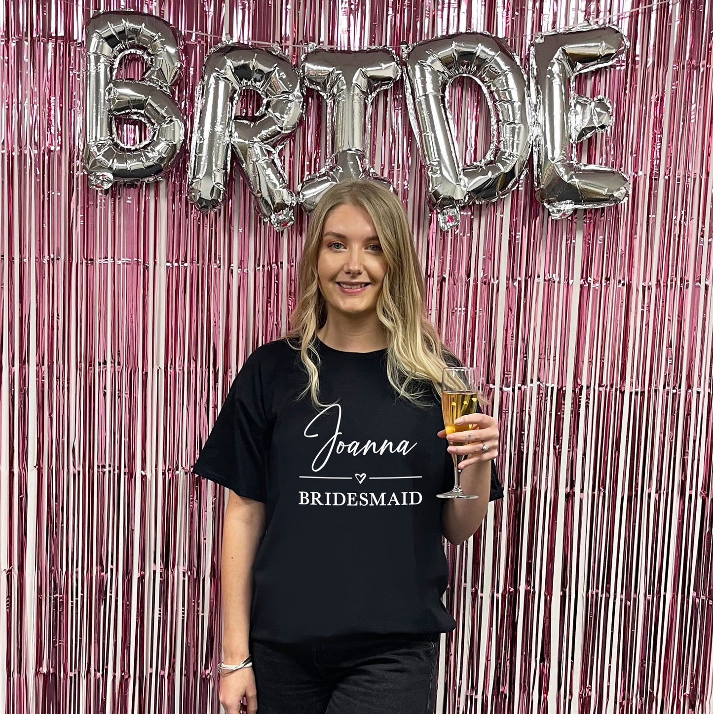Woman in a 'Joanna Bridesmaid' shirt holding a glass, with 'BRIDE' balloons in the background.