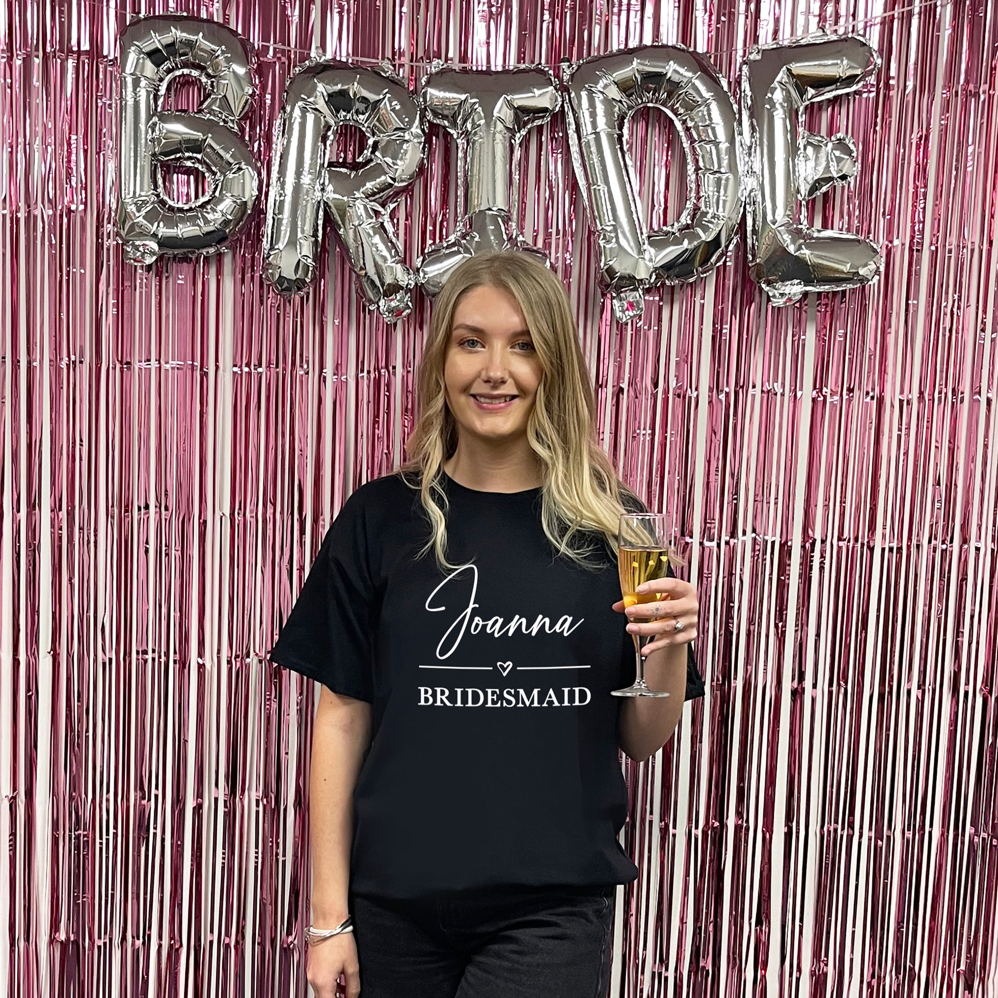 Woman in a 'Joanna Bridesmaid' shirt holding a glass, with 'BRIDE' balloons in the background.