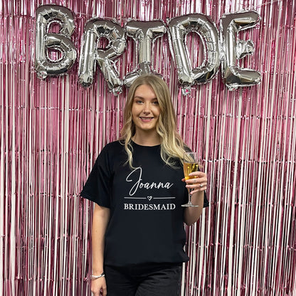 Woman in a 'Joanna Bridesmaid' shirt holding a glass, with 'BRIDE' balloons in the background.