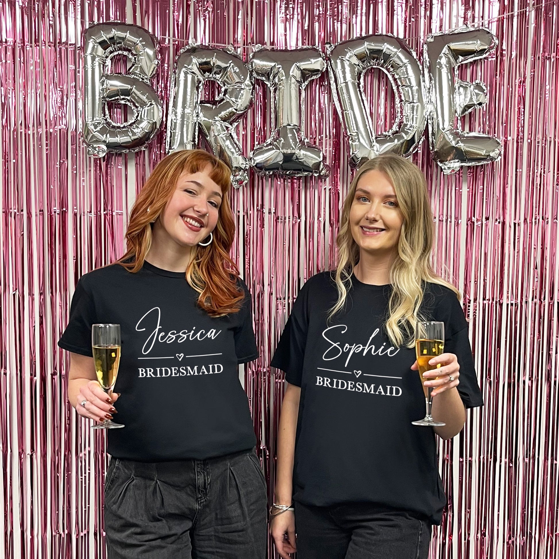 Two women wearing 'Bridesmaid' shirts with champagne glasses in front of a 'BRIDE' balloon setup.