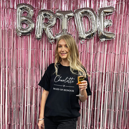 Woman in a 'Maid of Honour' shirt holding a glass, with 'BRIDE' balloons in the background.