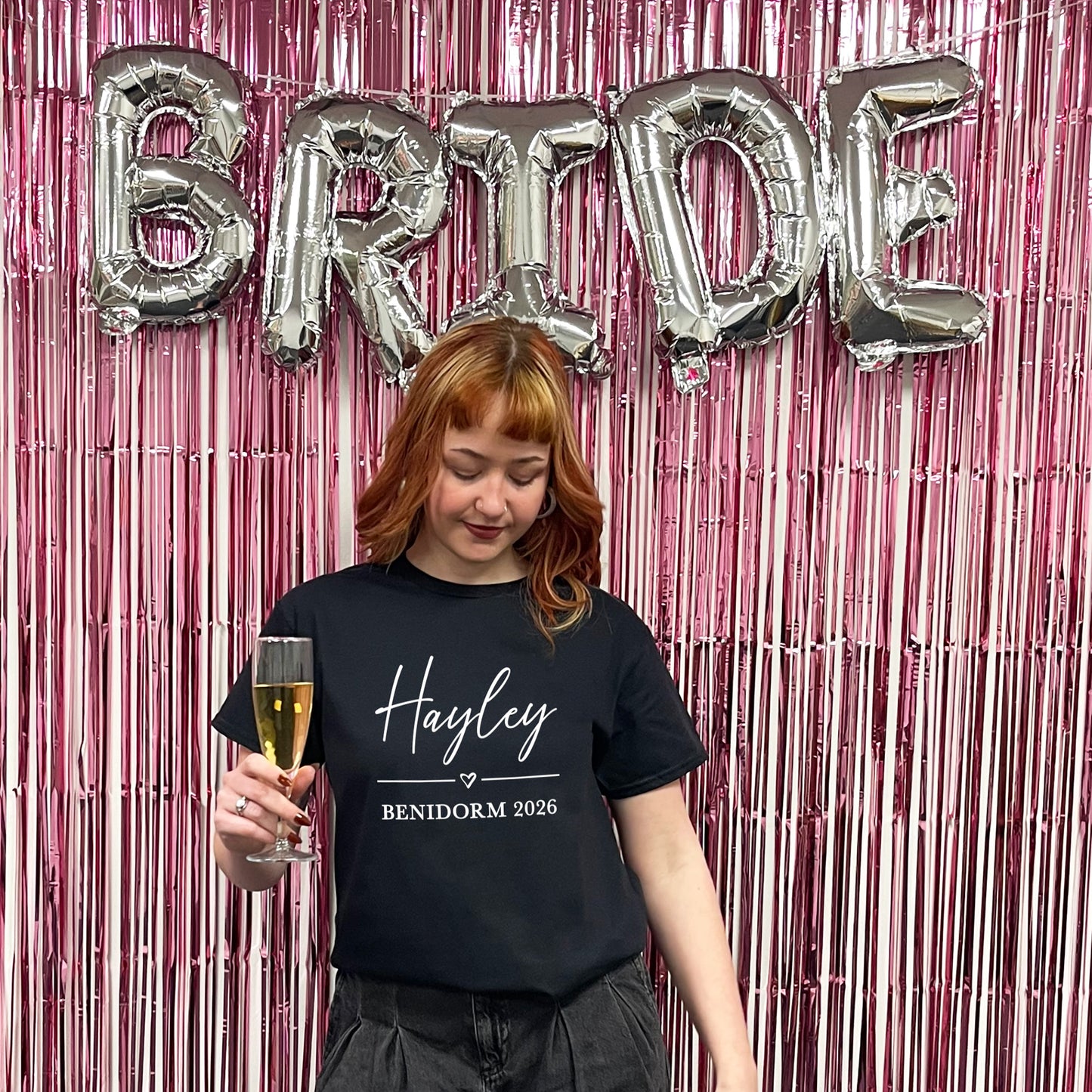Woman holding a glass of champagne with 'BRIDE' balloons and pink tinsel in the background