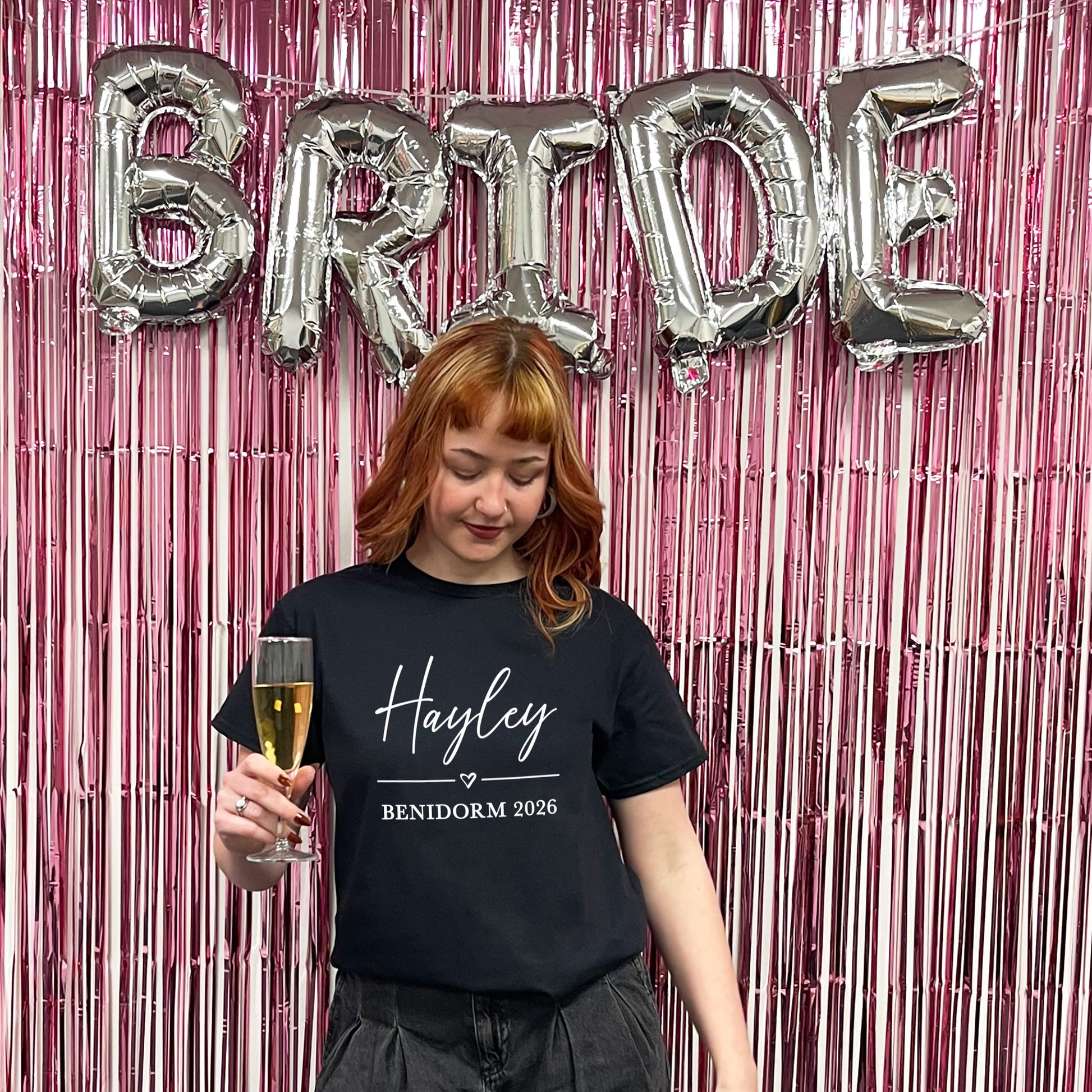 Woman holding a glass of champagne with 'BRIDE' balloons and pink tinsel in the background
