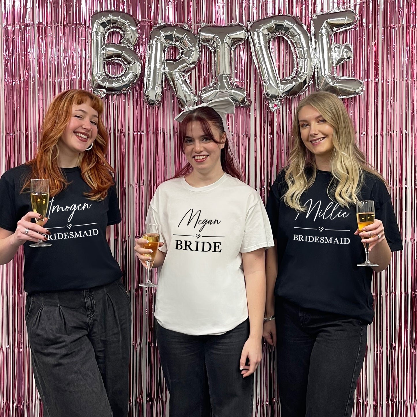 Three women posing with 'Bride' balloons and personalized shirts against a pink and silver decorative backdrop.