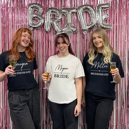 Three women posing with 'Bride' balloons and personalized shirts against a pink and silver decorative backdrop.