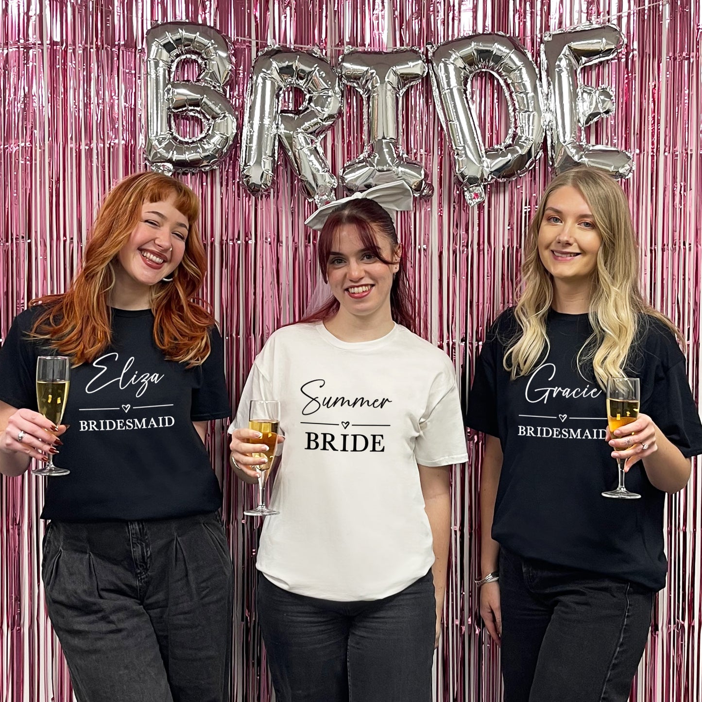 Three women in t-shirts with names on, holding drinks, in front of a 'BRIDE' balloon and pink curtain.
