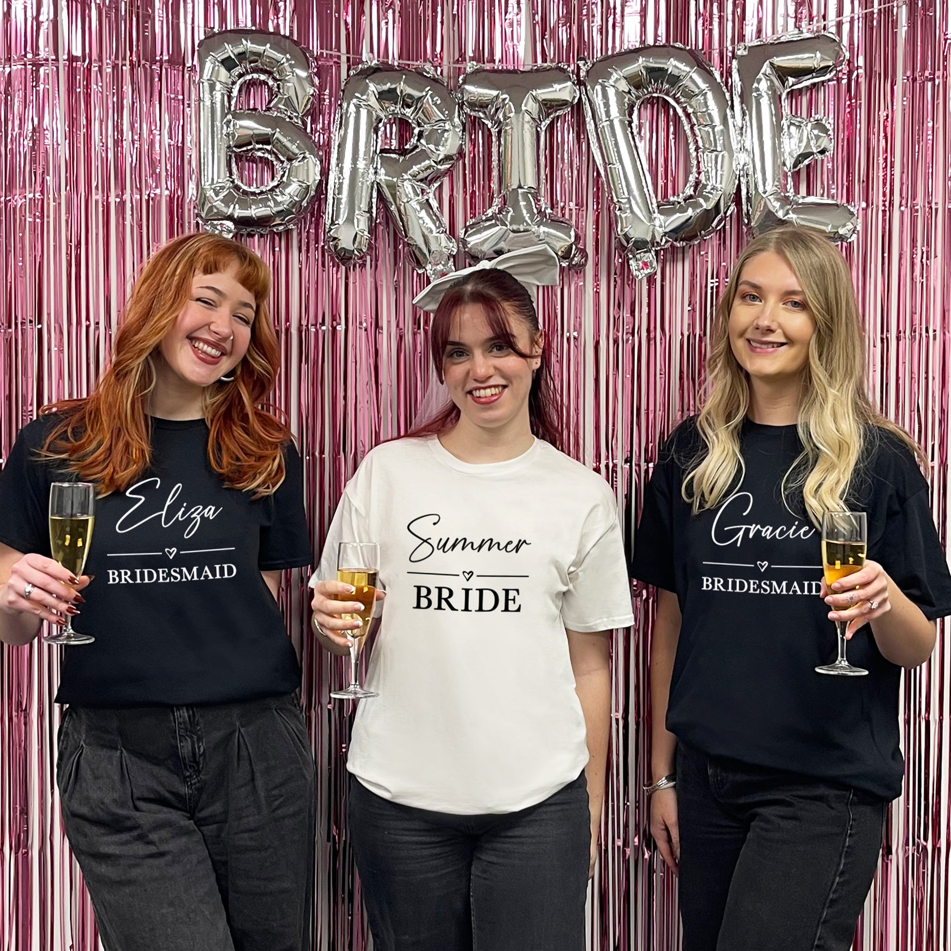 Three women in t-shirts with names on, holding drinks, in front of a 'BRIDE' balloon and pink curtain.