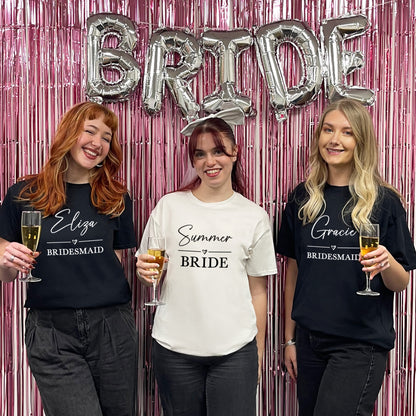 Three women in t-shirts with names on, holding drinks, in front of a 'BRIDE' balloon and pink curtain.