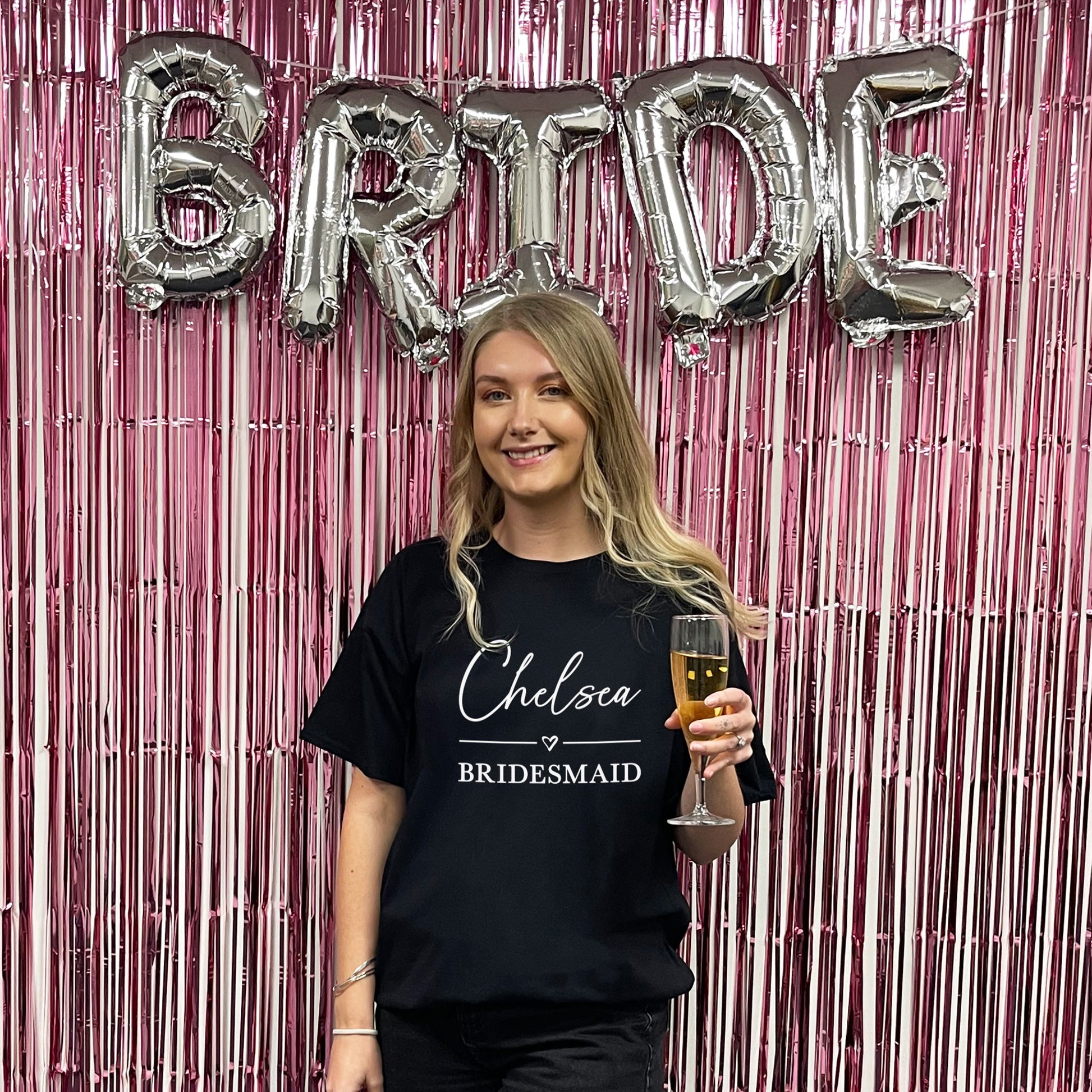 Woman in a 'Chelsea Bridesmaid' shirt holding a glass of champagne in front of 'BRIDE' balloons.
