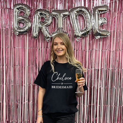 Woman in a 'Chelsea Bridesmaid' shirt holding a glass of champagne in front of 'BRIDE' balloons.