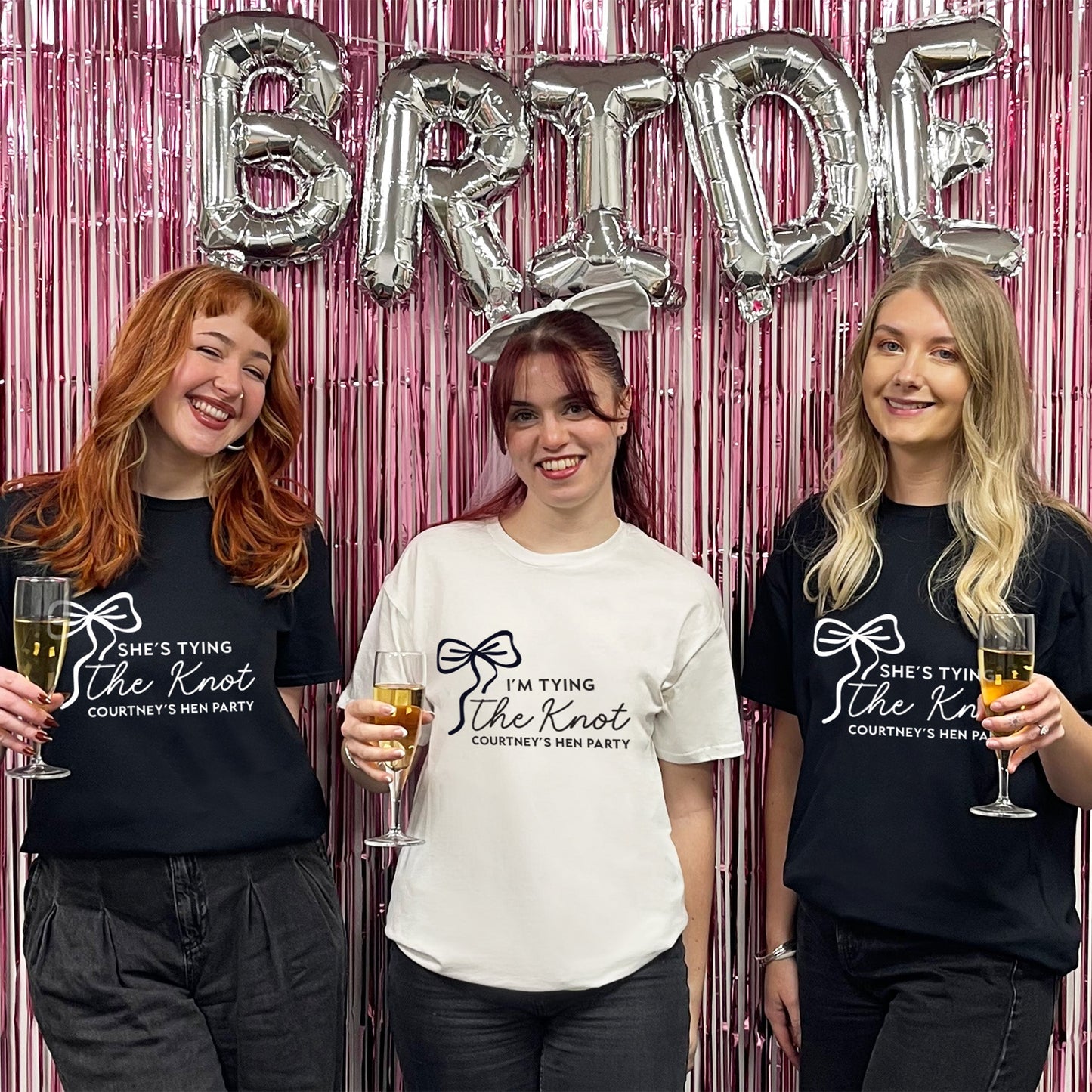 Three women posing with 'Bride' balloons and themed t-shirts against a pink metallic curtain.