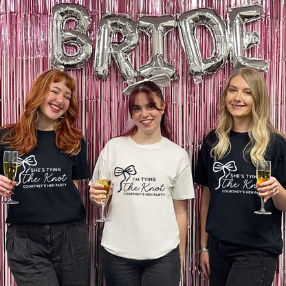 Three women posing with 'Bride' balloons and themed t-shirts against a pink metallic curtain.