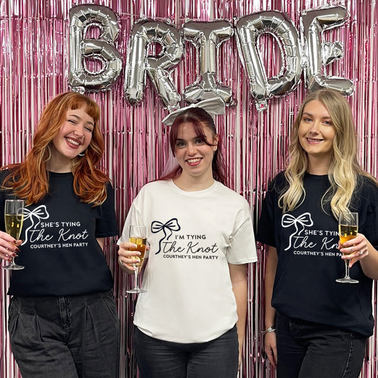 Three women posing with 'Bride' balloons and themed t-shirts against a pink metallic curtain.