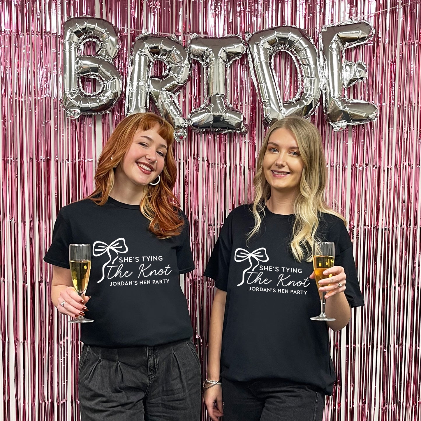 Two women wearing 'She's Tying the Knot' shirts with balloons spelling 'BRIDE' in the background.