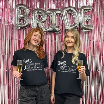 Two women wearing 'She's Tying the Knot' shirts with balloons spelling 'BRIDE' in the background.