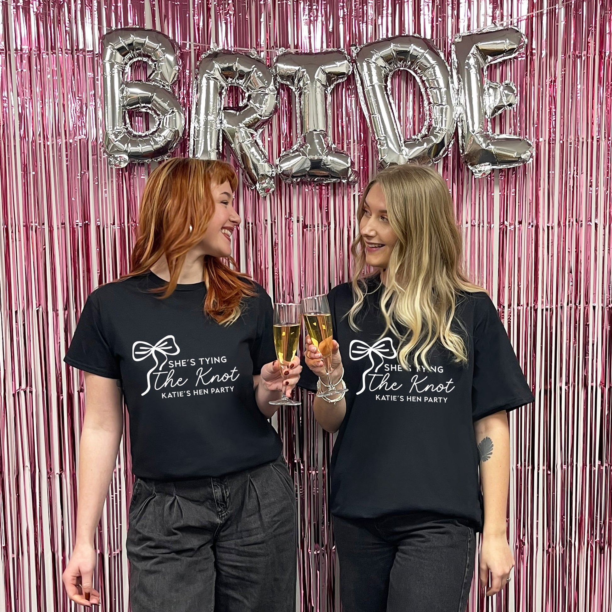 Two women wearing black t-shirts with 'She's Tying the Knot' text, holding drinks against a pink 'BRIDE' balloon backdrop.